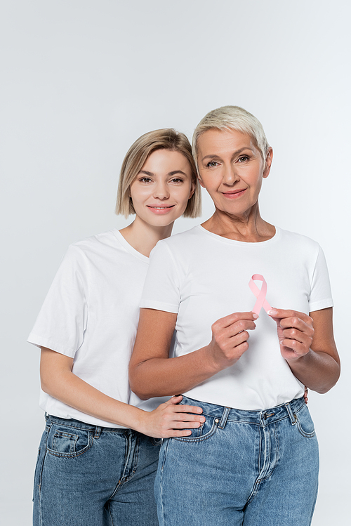 Smiling woman hugging senior friend with ribbon of breast cancer awareness isolated on grey