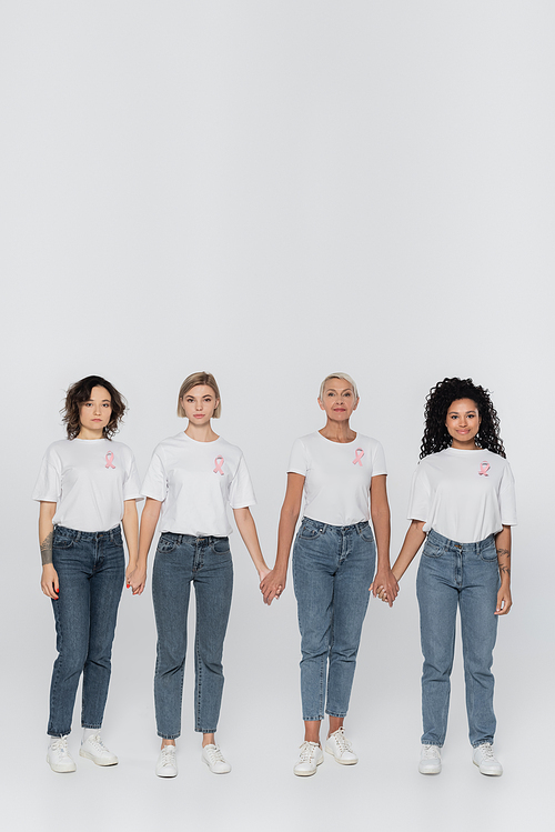 Multiethnic women with ribbons of breast cancer awareness holding hands on grey background