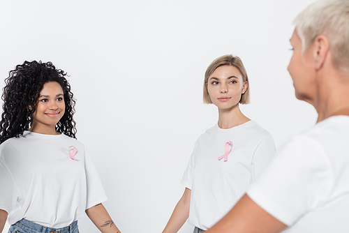 Smiling interracial women with ribbons of breast cancer awareness looking at senior friend isolated on grey