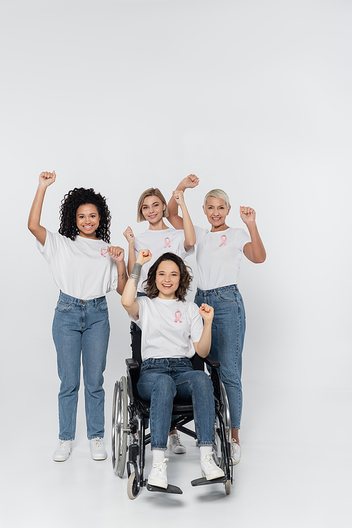 Smiling interracial women with pink ribbons showing yes gesture near friend in wheelchair on grey background