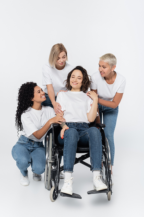 Smiling multiethnic women hugging friend in wheelchair on grey background