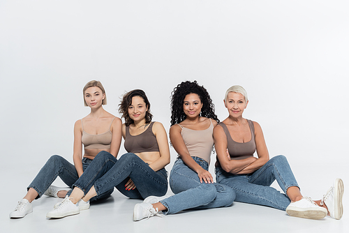 Positive interracial women smiling at camera while sitting on grey background