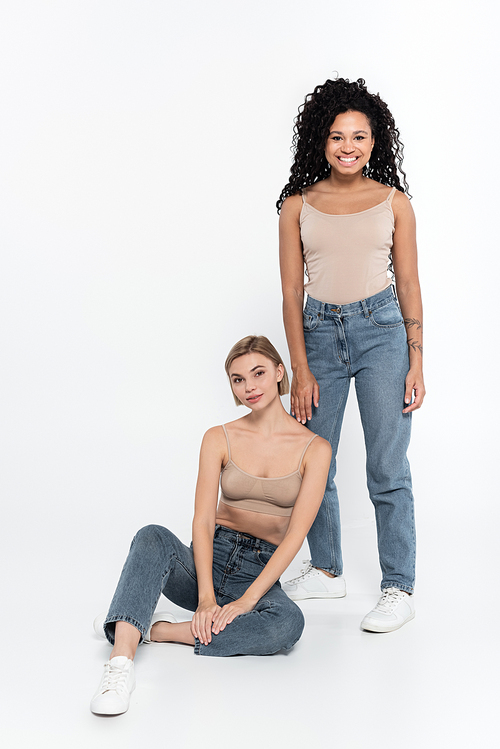 Cheerful african american woman in jeans standing near friend on grey background, feminism concept