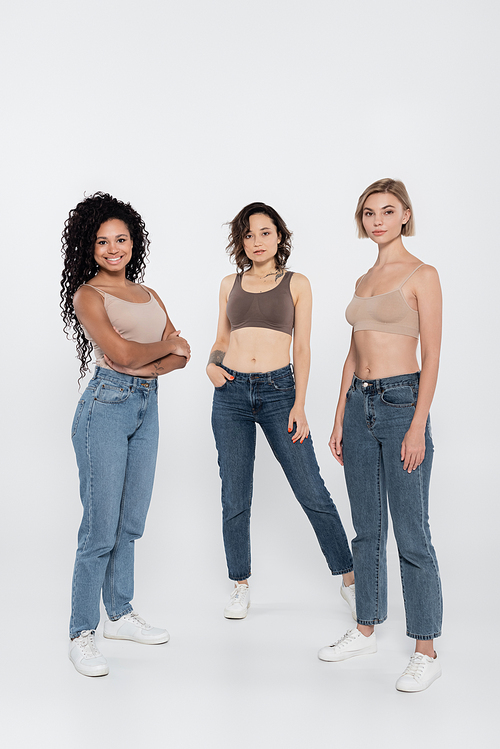 Young interracial women in tops and jeans posing on grey background
