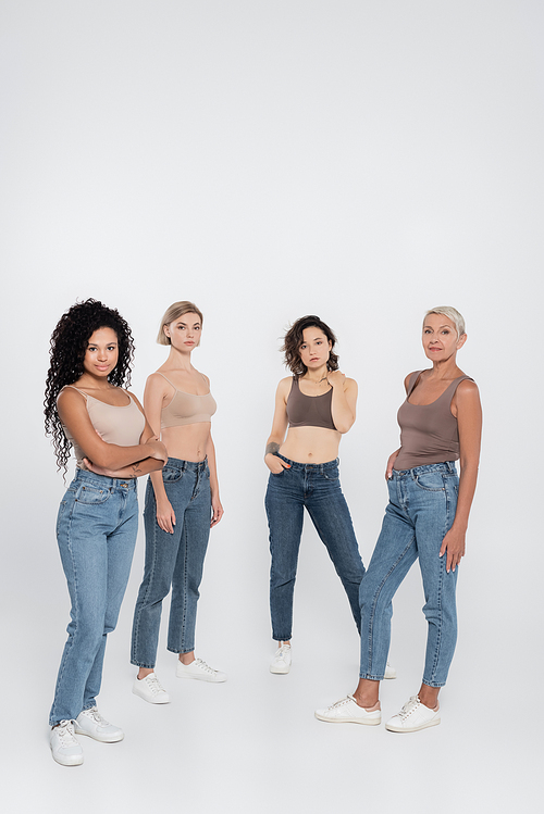 Multiethnic women in jeans posing and looking at camera on grey background