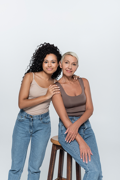 Curly african american woman hugging senior woman sitting on chair isolated on grey