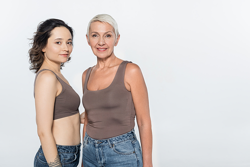 Brunette woman hugging cheerful elderly friend isolated on grey, feminism concept