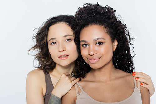 Young brunette woman hugging african american friend isolated on grey, feminism concept