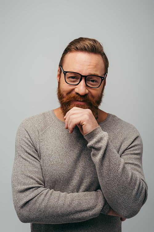 bearded man in long sleeve and eyeglasses smiling while looking at camera isolated on grey