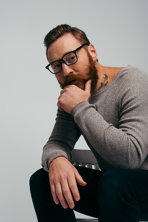 Portrait of bearded man in eyeglasses touching chin while sitting on chair isolated on grey