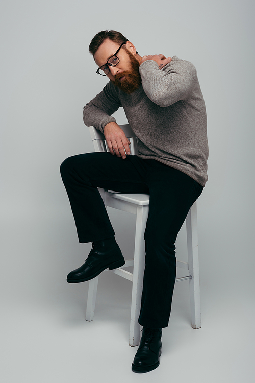 Full length of fashionable bearded man touching neck while sitting on chair on grey background