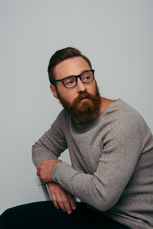 Fashionable model in eyeglasses and jumper sitting on chair isolated on grey