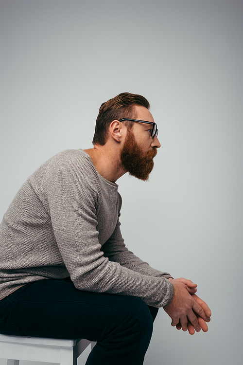Side view of bearded man in eyeglasses looking away while sitting on chair isolated on grey