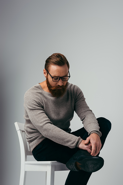 Trendy bearded man in eyeglasses touching leg while sitting on chair on grey background