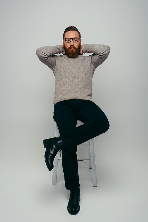 Full length of stylish man in eyeglasses touching neck and looking at camera while sitting on chair on grey background