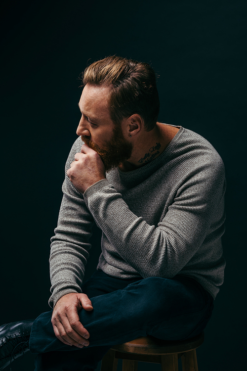 Side view of stylish tattooed man in grey jumper sitting on chair isolated on black