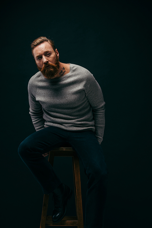 Bearded man in stylish grey jumper sitting on chair isolated on black