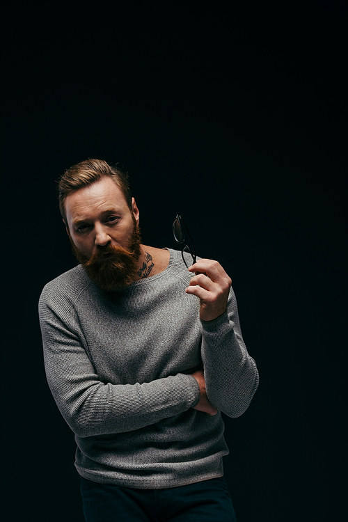 Stylish bearded man in jumper holding eyeglasses isolated on black with shadow