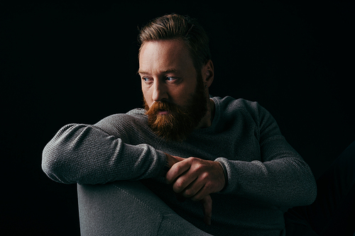 Bearded model in grey jumper looking away while sitting on armchair isolated on black