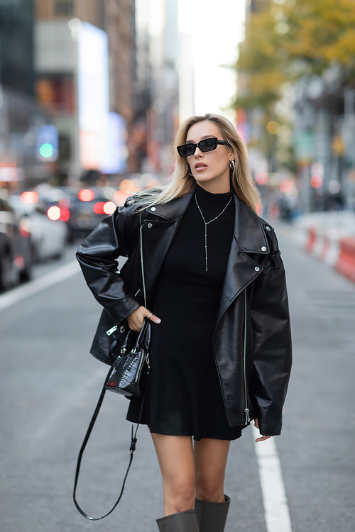 stylish woman in leather jacket and black dress standing with hand on hip on street of New York