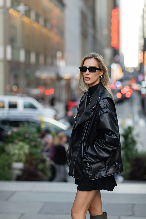 stylish woman in leather jacket and black dress standing with hand in pocket in New York