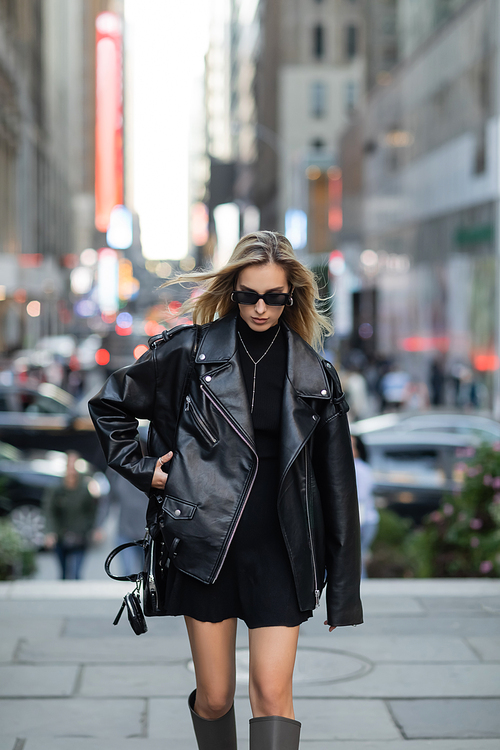 blonde woman in leather jacket and trendy sunglasses walking on street of New York