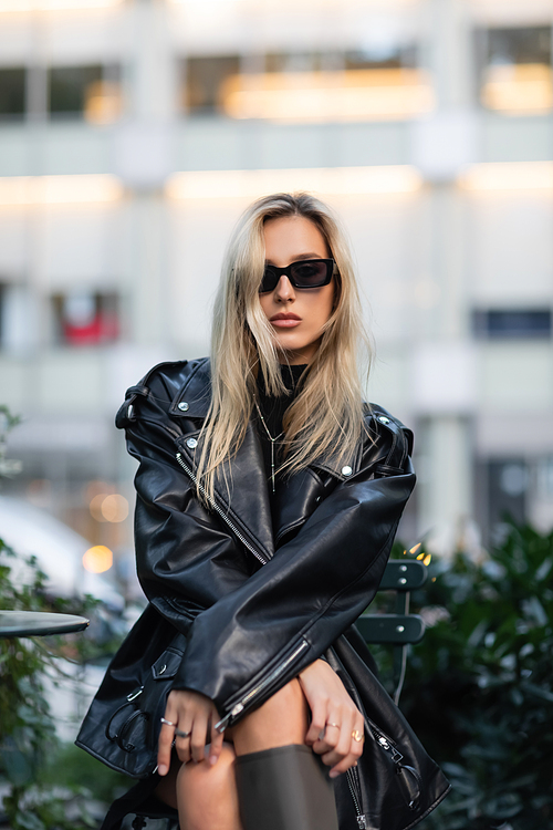 young blonde woman in black leather jacket and sunglasses sitting in outdoor cafe