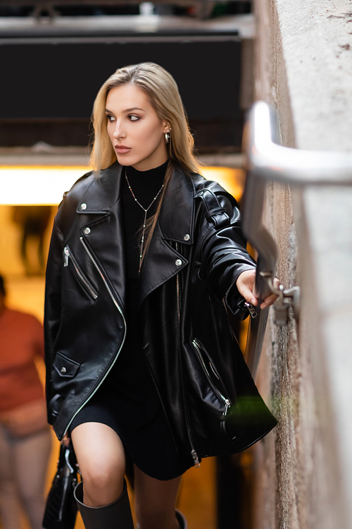 stylish young woman in leather jacket and black dress holding metal handrail near subway entrance in New York