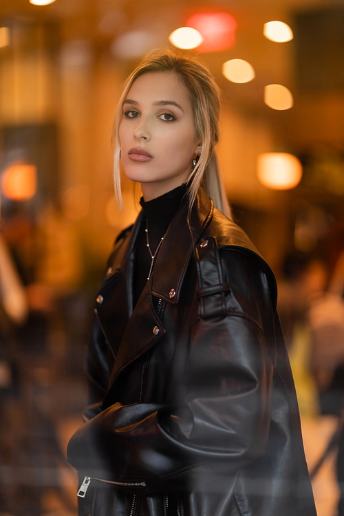 stylish woman in black leather jacket looking at camera through glass window in New York
