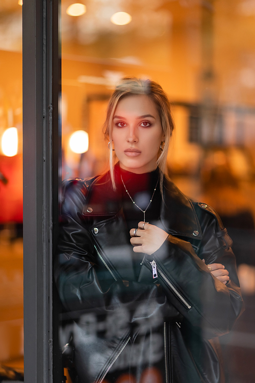 blonde young woman in black leather jacket looking at camera through glass window in New York