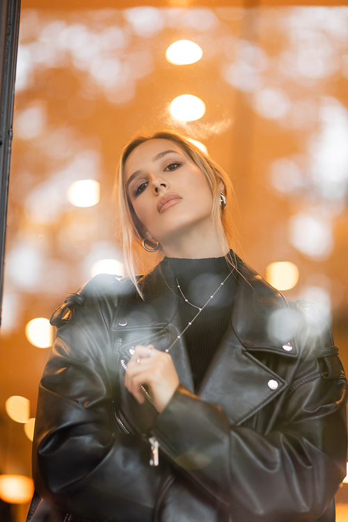 low angle view of stylish woman in black leather jacket looking at camera through glass window in New York
