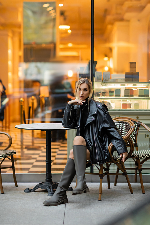full length of blonde woman in leather jacket sitting near round bistro table in outdoor cafe in New York