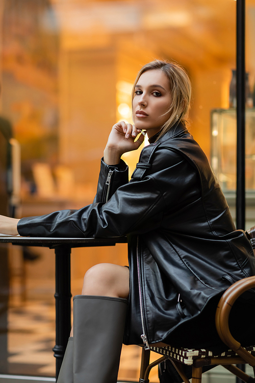 full length of young woman in black leather jacket sitting near round bistro table in outdoor cafe in New York