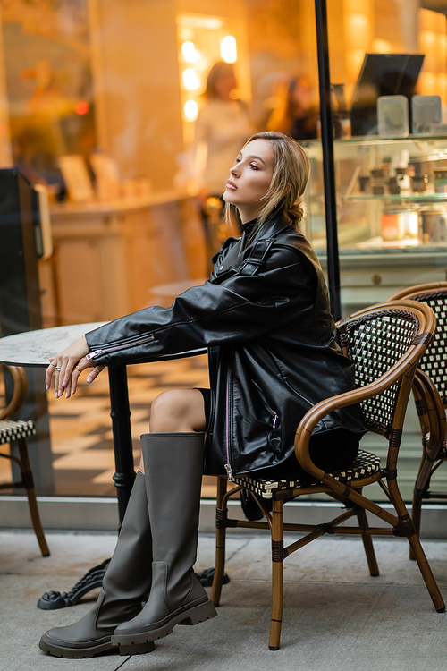 full length of stylish woman in black leather jacket and boots sitting near bistro table in outdoor cafe in New York