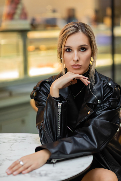 young blonde woman in black leather jacket sitting near bistro table in outdoor cafe in New York