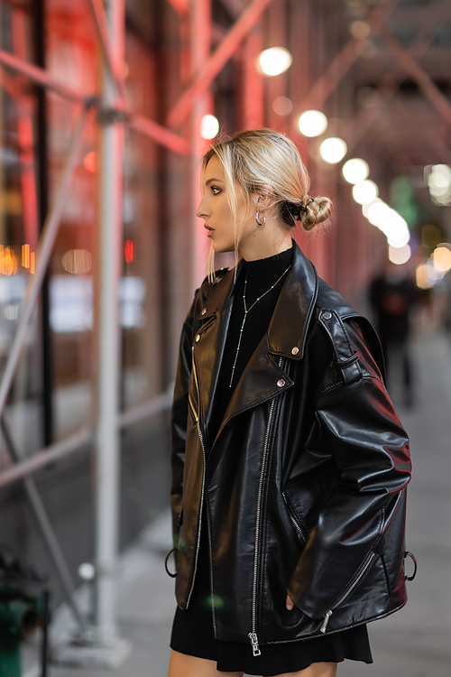 side view of blonde woman in black leather jacket standing on street in New York city at evening time