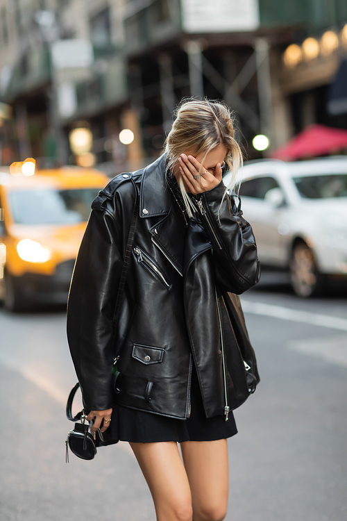blonde woman in black leather jacket and dress covering face with hand on street in New York