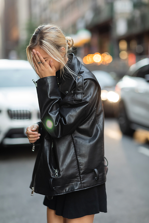 blonde woman in black leather jacket covering face with hand on street in New York