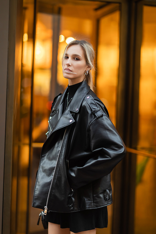 portrait of young and blonde woman in black leather jacket looking at camera while standing with hand in pocket outside