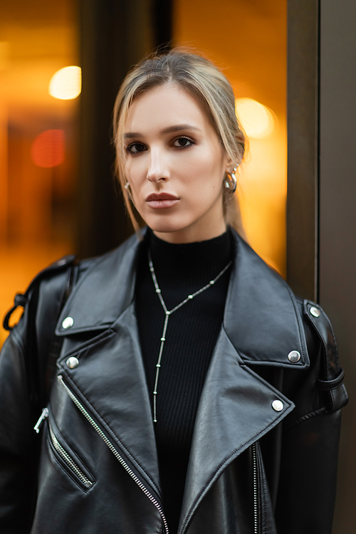 portrait of young and blonde woman in black leather jacket looking at camera outside