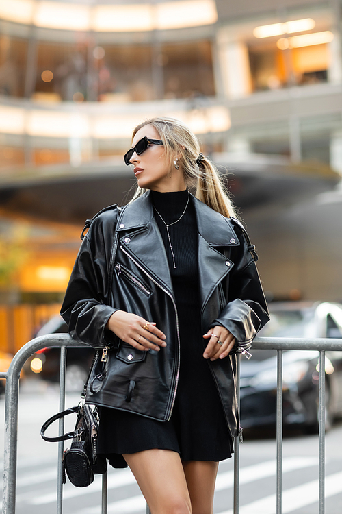 stylish woman in leather jacket and dress standing with handbag near metal barrier in New York