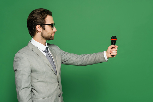 Side view of newscaster in suit holding microphone and looking away on green background