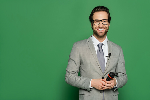 cheerful news anchor in suit and eyeglasses holding microphone on green background