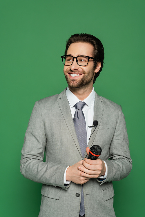 happy news anchor in suit and eyeglasses holding microphone isolated on green