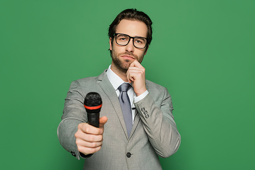 pensive news anchor in suit and eyeglasses holding microphone isolated on green