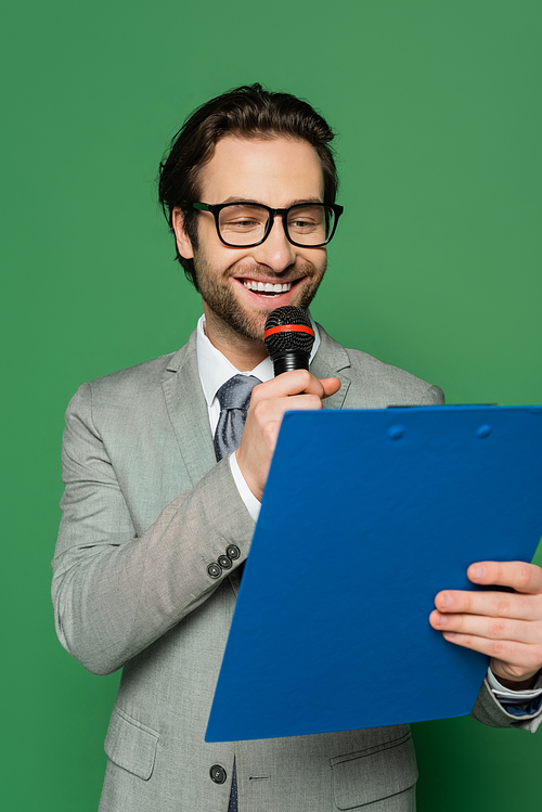 happy news anchor in suit and eyeglasses talking in microphone while holding clipboard isolated on green