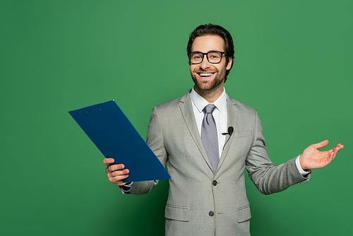 happy news anchor in suit and eyeglasses holding clipboard while gesturing on green background