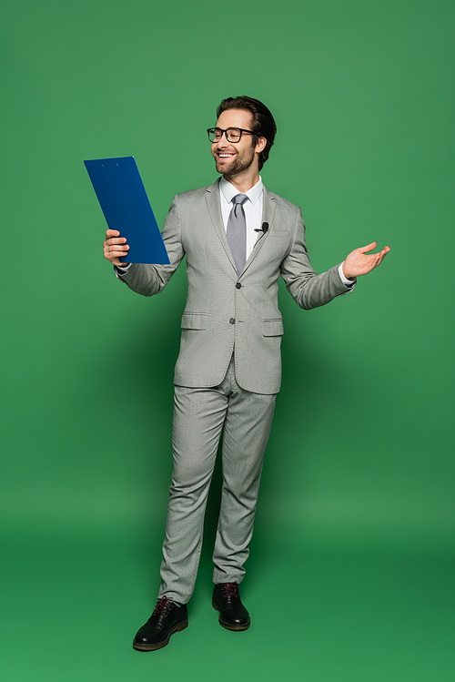 full length of cheerful news anchor in suit and eyeglasses holding clipboard while gesturing on green