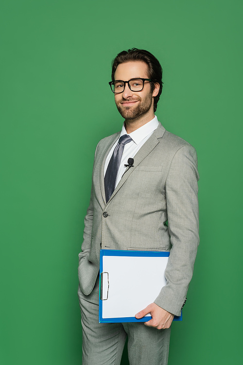 happy news anchor in suit and eyeglasses holding clipboard while standing with hand in pocket isolated on green