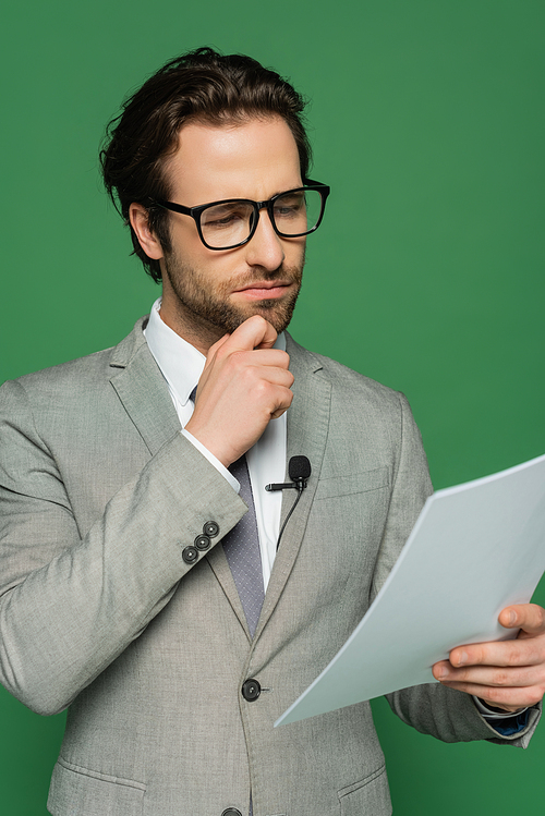 thoughtful news anchor in eyeglasses and suit looking at blank paper isolated on green